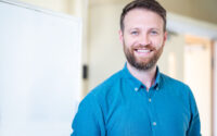 Smiling tutor in a blue shirt standing by a whiteboard.
