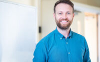 Smiling tutor in a blue shirt standing beside a whiteboard.