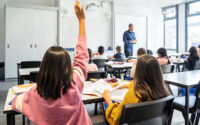 Pupil raising her hand while the tutor stands at the front of the classroom.