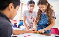 Two girls working on a hands-on activity at a desk, watched by a classmate.