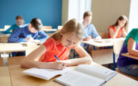 Girl writing in her exercise book with a textbook open, while classmates work behind her.