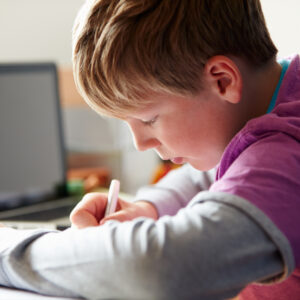 Boy concentrating as he writes at a desk, with a laptop behind him.